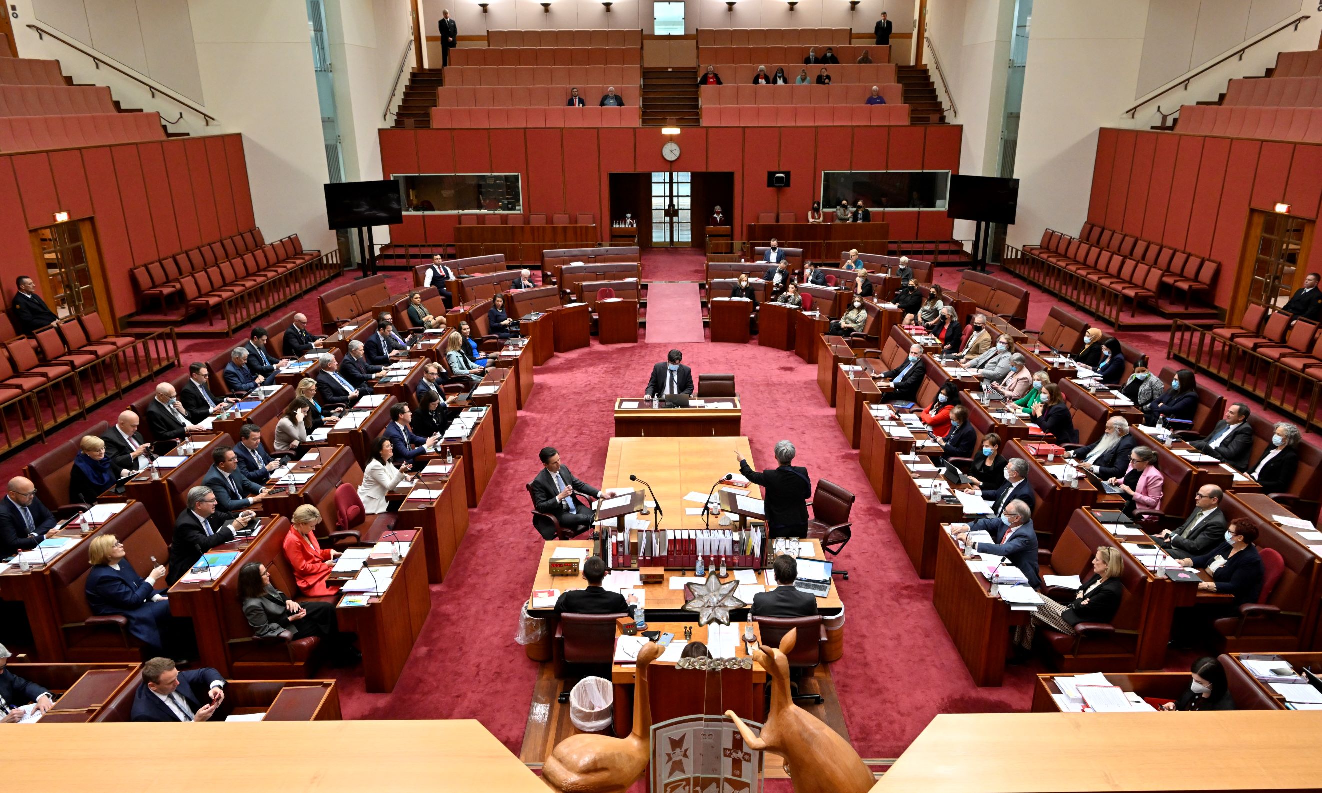 The red Senate chamber. There are people sitting in seats which are arranged in a U-shape around a large central table. 