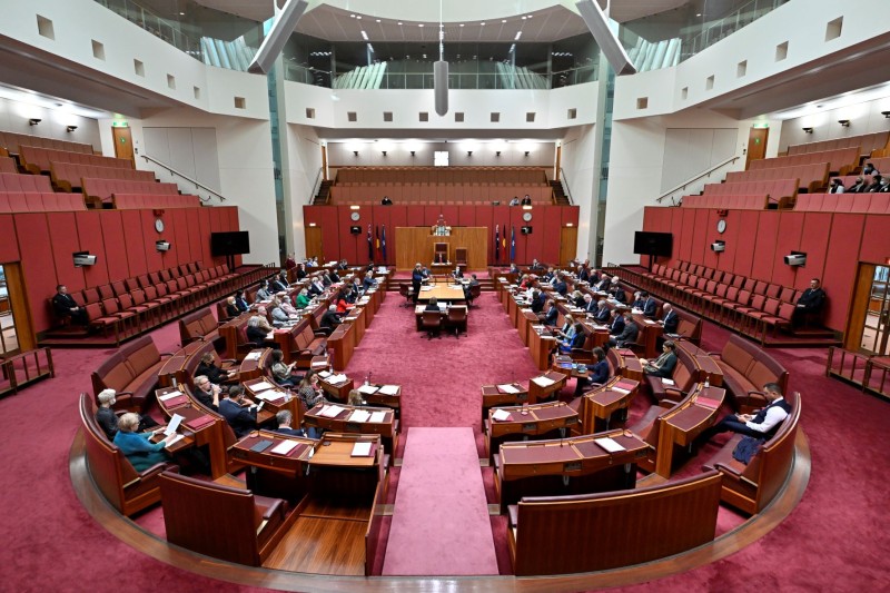 The red Senate chamber. There are people sitting in seats which are arranged in a U-shape around a large central table. 