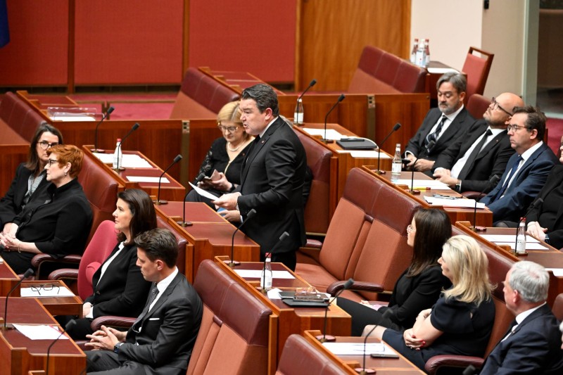 A man in a dark suit is standing at a desk in a red room. He is speaking. Other men and women also in suits sit at desks around him.